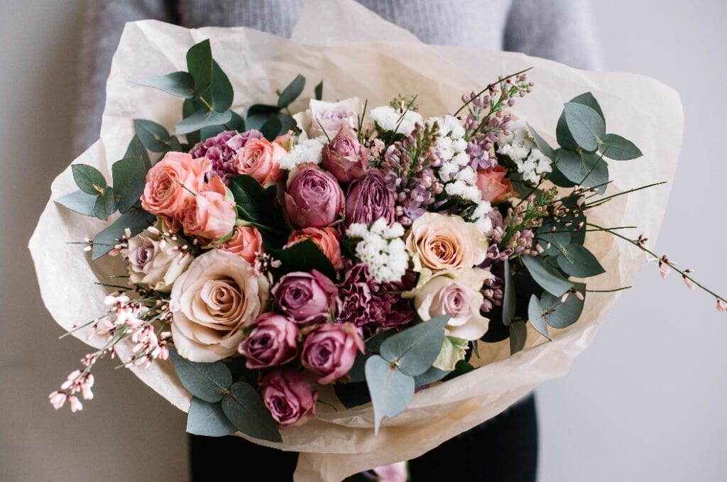 Young florist woman holding freshly made blossoming flower bouquet of roses and eucalyptus on the grey wall background.