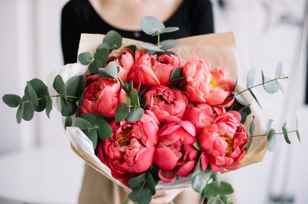 Very nice young woman holding beautiful tender blossoming mono bouquet of fresh coral peony flowers and eucalyptus on the rustic wooden wall background